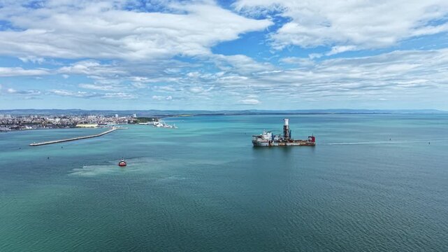 An industrial dredging vessel operates near the coast close to a busy port. The sun shines above, and a small boat is visible in the water nearby.