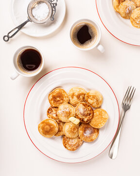 Dutch poffertjes, small pancakes, are arranged on white plates with a pat of butter and powdered sugar, accompanied by coffee cups. View from above.