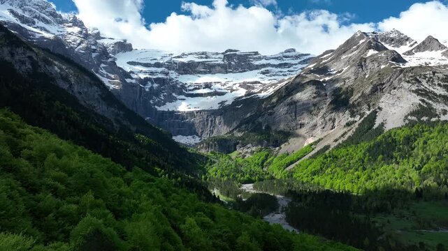 Snowy walls of the Cirque de Gavarnie rising above lush green forested valley under blue sky