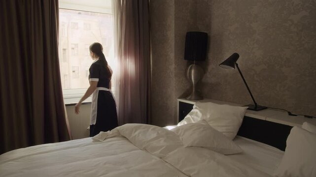 Wide shot of young adult Caucasian woman in maid uniform opening window curtains and making bed in hotel room. Footage ideal for hospitality industry marketing and hotel service promotions