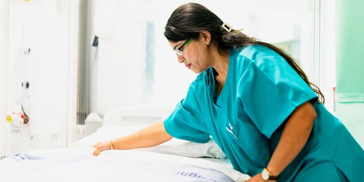 Nurse in teal scrubs tending to a hospital bed in a bright room.