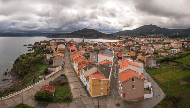 Aerial view of the coastal town of Carino with its colorful houses and orange rooftops situated along the rugged coastline under a dramatic cloudy sky Carino, A Coruna, Spain.