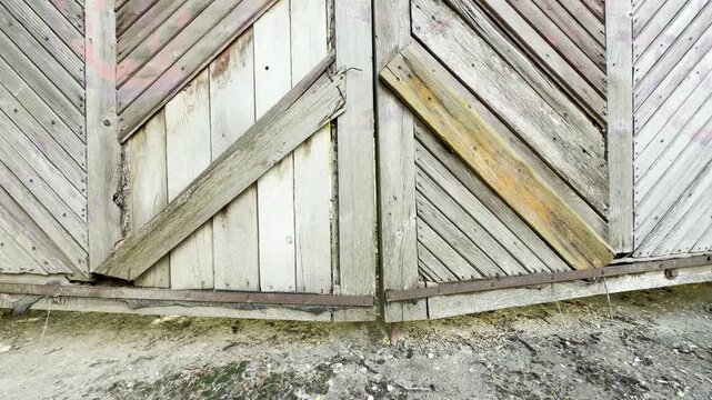 Large weathered wooden gates stand closed together. The textured timber planks block an entrance over dry dirt ground.