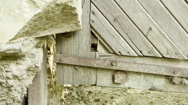 A weathered wooden gate meets a rough stone wall. The old timber structure with a rusty metal bar hangs over dry dirt ground in a rustic courtyard.