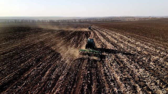 Agricultural machinery plowing soil in a large field, with multiple tractors visible working in unison, creating dust clouds in the rural landscape under clear skies