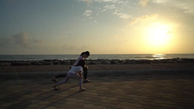 Healthy mother and her young daughter doing side lunge stretching exercises together on a coastal brick path during a beautiful golden sunrise by the sea
