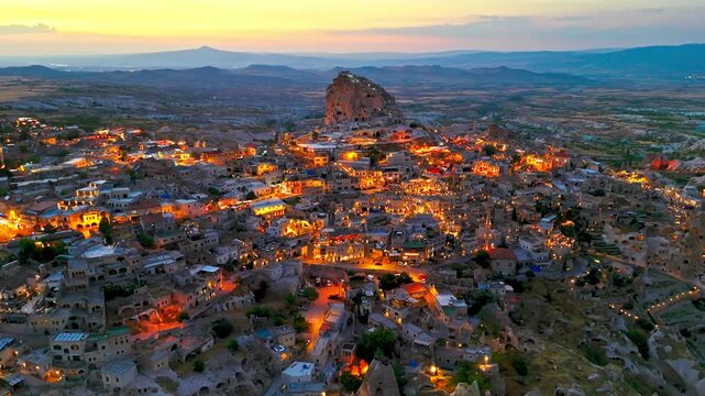 Drone view of Uchisar, Cappadocia, Turkey at sunset. Golden sky, glowing rooftops and Uchisar Castle shape a scenic evening panorama.