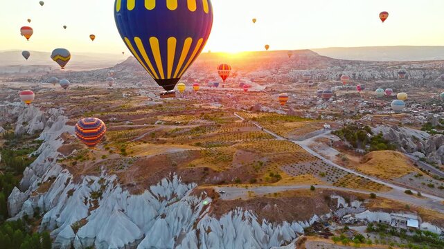 Aerial view of Rose Valley, Cappadocia, Turkey at sunrise. Hot air balloons drift across the sky above rugged cliffs and glowing valley terrain.