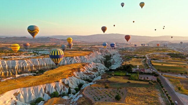 Aerial view of Rose Valley, Cappadocia, Turkey at sunrise. Balloons float above the road, rocky hills and sunlit open terrain.
