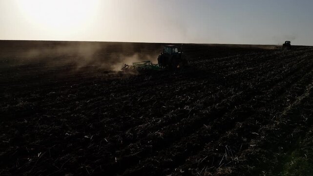 Tractors plowing a field under the sun, creating dust clouds while preparing the soil for planting in a rural agricultural landscape