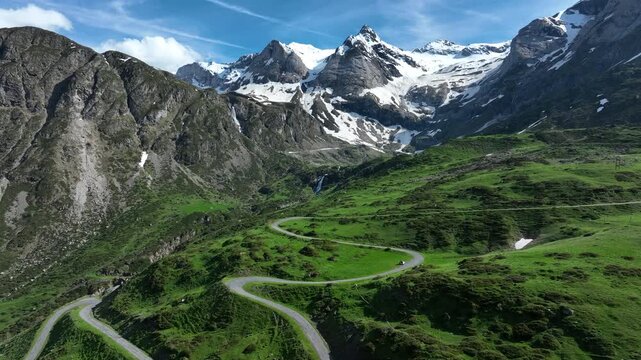 Aerial view of winding hairpin mountain road through green meadow below snowy Pyrenees peaks