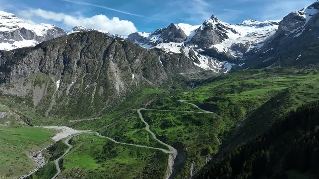 Aerial view of winding mountain road through green valley below snowy jagged Pyrenees peaks