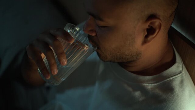 Man taking sleeping pills before bedtime while sitting on bed at home. Sleep aid medication, nighttime routine, stress management, healthcare and wellness concept.
