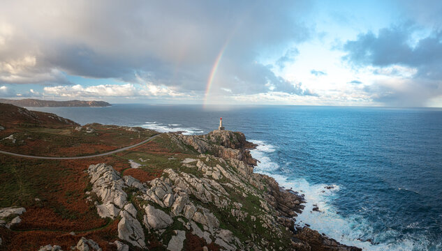 Aerial view of Faro de Punta Nariga with a rainbow arching over the lighthouse, rugged rocky cliffs, and the Atlantic Ocean under a cloudy sky Malpica, A Coruna, Spain.