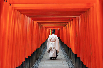 Fototapeta premium Traditional Shinto priest walking alone from behind through the vibrant orange Torii gates at Fushimi Inari Shrine in Kyoto, Japan on an early morning