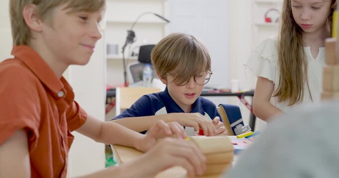 Young boy with glasses builds wooden block structure while classmates join table activity, creating primary classroom teamwork, peer interaction, confidence, and back to school engagement today.