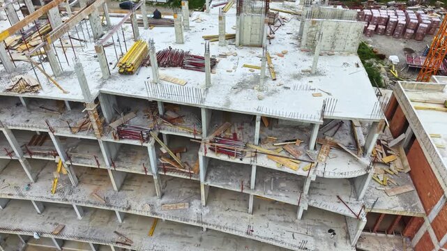 Aerial view curved concrete building under construction with stacked open floors, scaffolding, crews installing formwork and yellow hoists, dusty atmosphere,