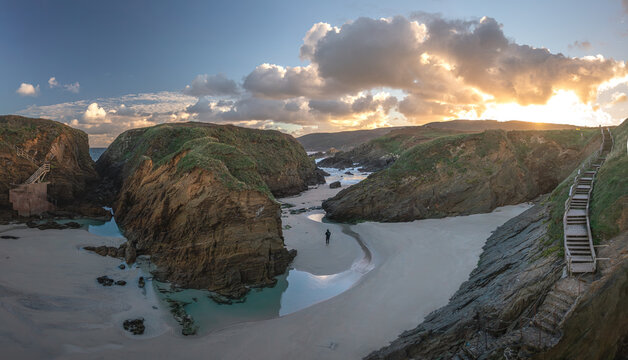 Aerial view of the rugged cliffs and sandy beach of Santa Comba with a wooden staircase and a person walking on the shore during sunset Covas, La Coruna, Spain.