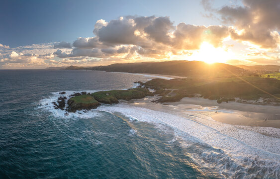 Aerial view of Ermita de Santa Comba on a rocky island during a golden sunset with waves breaking on the sandy beach and green coastal hills Covas, La Coruna, Spain.