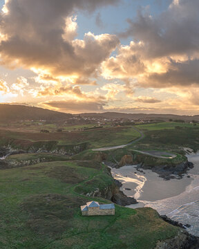 Aerial view of Ermita de Santa Comba, a small stone chapel on a green cliff overlooking the beach and ocean under dramatic sunset clouds in Covas, La Coruna, Spain.