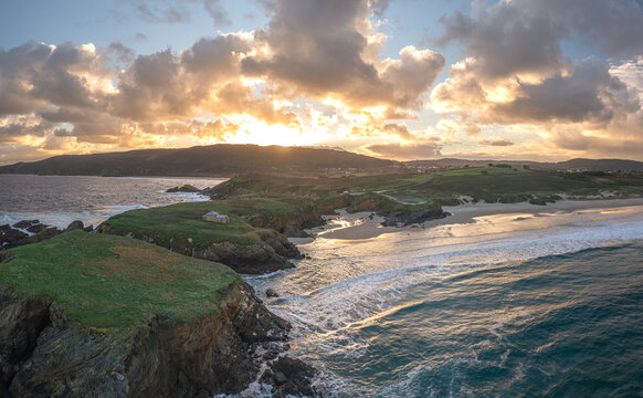 Aerial view of Ermita de Santa Comba on a rocky islet overlooking a sandy beach and crashing waves under a golden sunset sky in Covas, La Coruna, Spain.