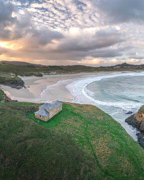 Aerial view of Ermita de Santa Comba on a green cliff overlooking the sandy beach and crashing waves under a dramatic sunset sky Covas, La Coruna, Spain.