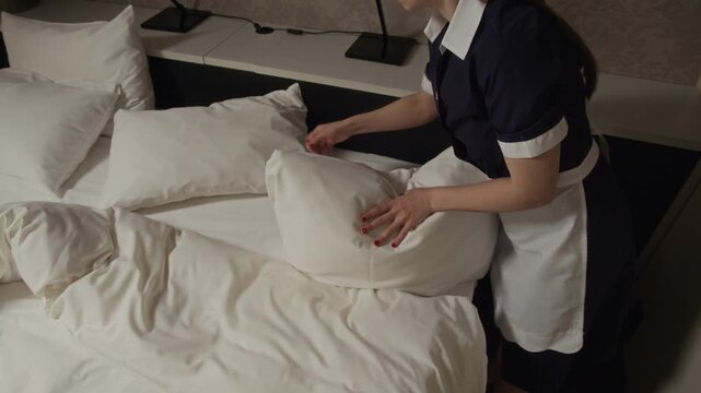 High angle view of young adult Caucasian woman in maid uniform making bed in hotel room, arranging pillows. Footage suits hospitality industry marketing and hotel service promotion