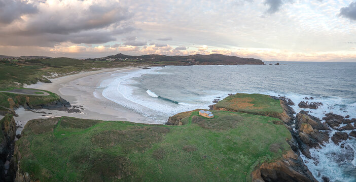 Aerial view of Ermita de Santa Comba on a green islet overlooking the sandy beach and Atlantic ocean waves in Covas, La Coruna, Spain.
