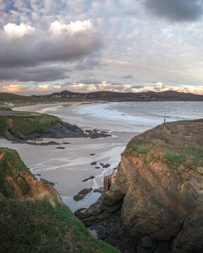 Aerial view of Ermita de Santa Comba on a rocky island overlooking the sandy beach and crashing waves under a cloudy sky in Covas, La Coruna, Spain.