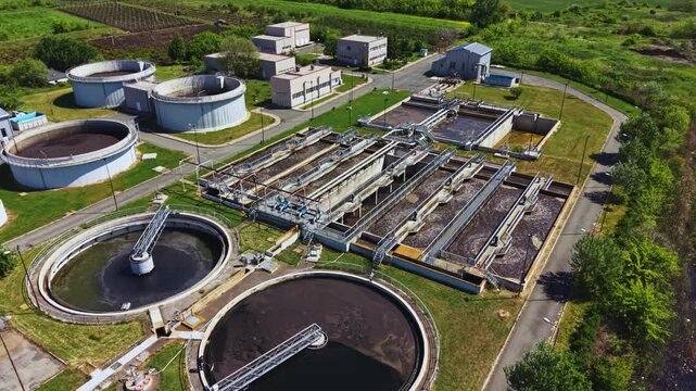 Aerial sewage treatment plant showing circular clarifiers and aeration basins, sludge tanks, pipe networks, control buildings, green buffer vegetation, slow