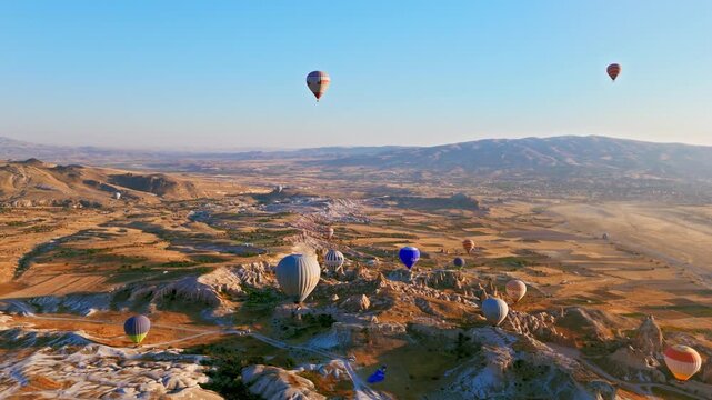 Drone view of Love Valley, Cappadocia, Turkey. Multiple balloons float above fairy chimneys, pale rocks and blue morning sky.