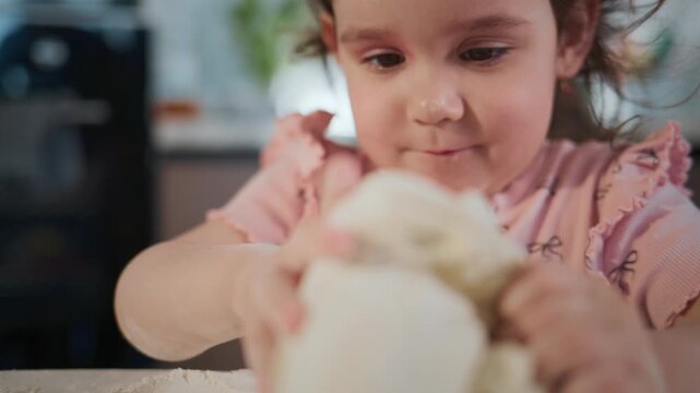 Young girl kneads dough with focused determination. Her hands press and stretch the dough with care. She wears a pink dress with bow patterns. Her expression shows concentration and playful enjoyment