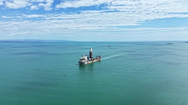 An industrial dredging vessel moves through clear waters in a coastal area. The ocean reflects light, and far off, other ships can be seen on the horizon.