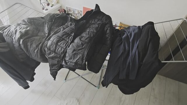Freshly washed dark clothes, including down jackets and shirts, hanging on a portable drying rack inside a house with wooden flooring