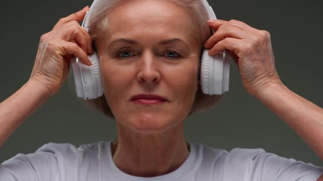 Portrait: happy female pensioner puts headsets on head. Face of senior old woman with gray hair elderly grandmother. Listening to music or podcast with white headphones. Wrinkles on face, good mood