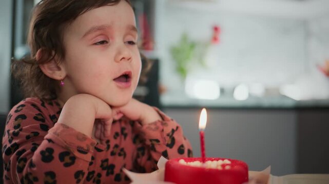 Little girl blows out a birthday candle. She closes her eyes, focused and calm. Her cheeks puff as she breathes out. Her hands rest under her chin, still and patient. She wears a patterned top, soft