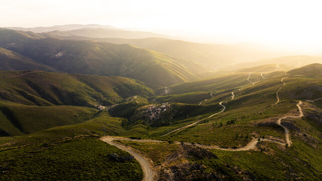 Aerial view of winding dirt roads snaking through lush green mountains and small villages during a golden sunset Aldeia das Dez, Coimbra, Portugal.