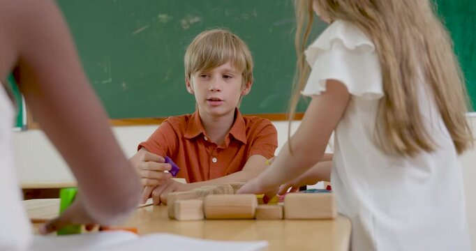Young blond boy sits at desk with girl classmates, stacking wooden blocks near chalkboard, building creativity, classroom participation, friendship, and back to school learning confidence today.