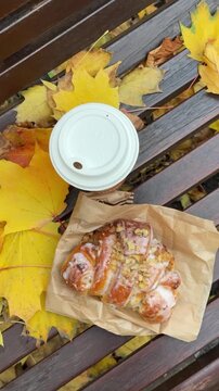 Paper coffee cup with hot drink and fresh croissant from bakery on bench covered with yellow fallen leaves from trees in autumn top view. Warming romantic coffee break in city park