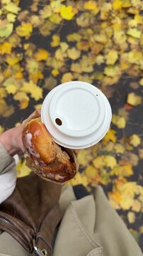 Woman hand with fresh pastry from bakery and hot coffee to go in paper cup with the background of ground fully covered with many fallen yellow leaves. Romantic walk in autumn park and coffee break