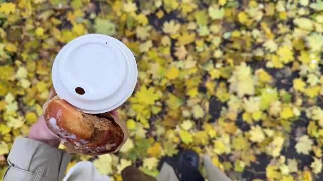 Woman hand with fresh pastry from bakery and hot coffee to go in paper cup with the background of ground fully covered with many fallen yellow leaves. Romantic walk in autumn park and coffee break