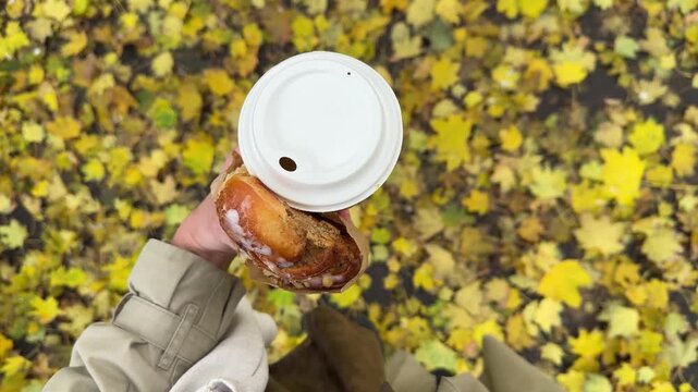 Woman in trench coat keeping in hand big paper coffee cup and fresh croissant from bakery and walking in autumn city park with ground covered with golden fallen leaves high angle view