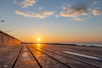 Fototapeta premium Golden morning sunrise over calm sea from low angle perspective on wooden boardwalk planks in off-season. Beautiful coastal landscape reflection. Ayia Napa, Cyprus.