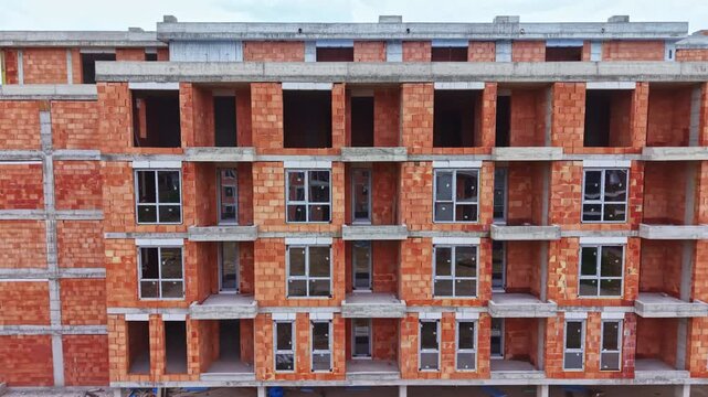 Unfinished brick apartment building facade showing exposed red brick and concrete slabs, empty window openings and balcony recesses, raw industrial atmosphere