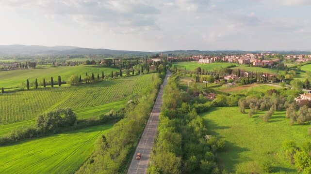 A stationary drone lift above a rural road in San Rocco a Pilli, Italy, showing a passing car, cypress trees, and the surrounding Tuscan landscape in soft afternoon light.