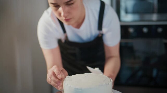 Baker carefully applies frosting to a cake on a turntable. She focuses intently, using a spatula for smooth layers. Kitchen tools and appliances surround her workspace. Ideal for bakery promotions