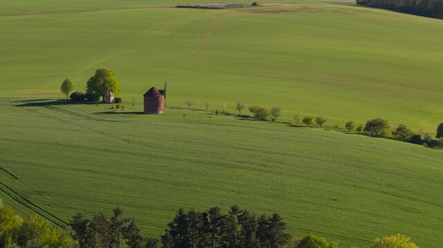 Aerial view of lush green agricultural field with old traditional windmill and scattered trees. Expansive countryside landscape with field divisions. Rural farming heritage in temperate climate.