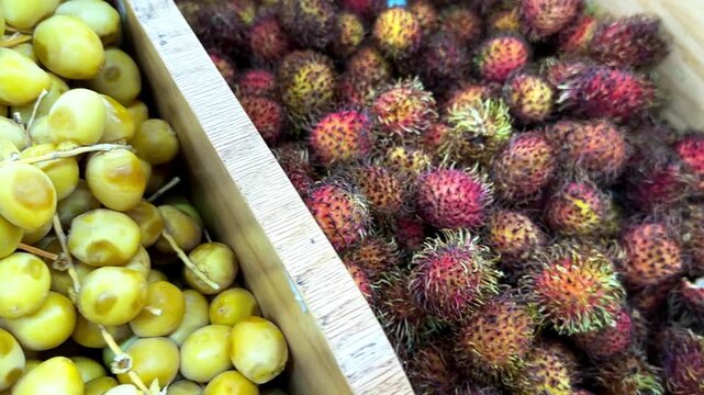 Close-up of a person's hand choosing a ripe, hairy rambutan from a wooden crate at a grocery store or farmer's market in slow motion