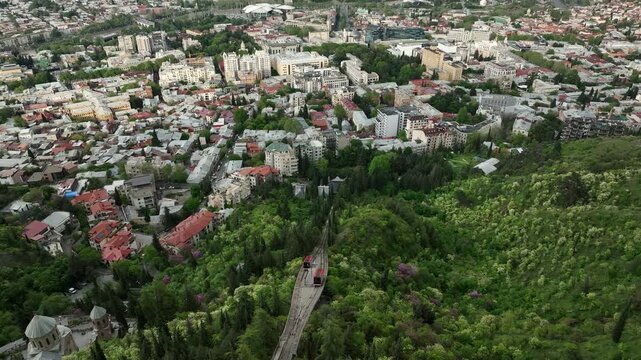 High angle aerial view of Tbilisi funicular tram cars passing each other on Mtatsminda hill, red railway cabins surrounded by green forest and city residential area, Georgia cityscape.