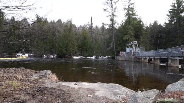 Landscape shot of a river flowing toward a small hydroelectric dam structure, framed by coniferous forest, remnants of snow, and a yellow floating containment boom in the water.